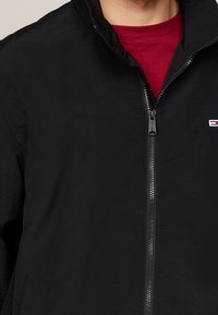 Zwarte zip-up jas met een gladde, lichte textuur, staande kraag en een klein logo op de linkerkant van de borst. De binnenvoering is rood.