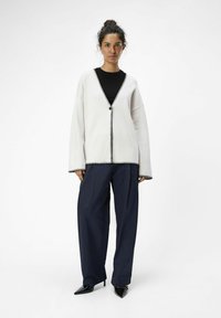 Lys grå cardigan med sort kant, oversized pasform, dyb V-hals og enkelt knaplukning, parret med marineblå wide-leg bukser og sorte hæle.