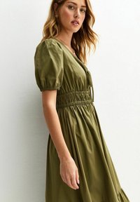 Vestido verde olivo con mangas abullonadas y cintura fruncida. La tela es suave y el diseño fluye de manera suelta por debajo de la cintura.