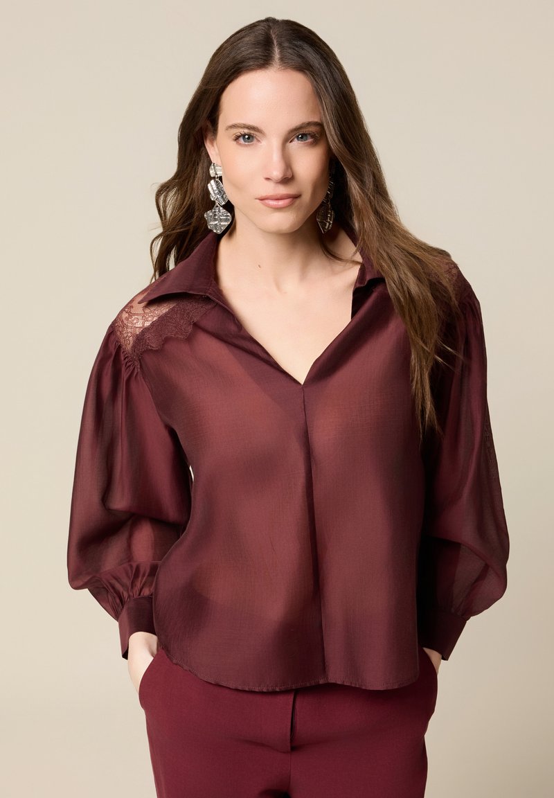 Blusa burdeos semitransparente con acentos de encaje, mangas largas abullonadas y escote en V; combinada con pantalones burdeos a juego.
