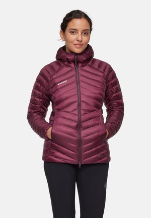 Vrouw die een bordeauxrode gewatteerde jas met rits draagt, met handen in de zakken, staand tegen een effen witte achtergrond.