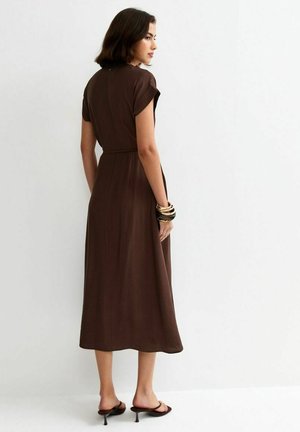 Vrouw die zijwaarts staat, gekleed in een donkerbruine jurk tot op de knie, zwarte sandalen met hak en meerdere armbanden aan één pols.