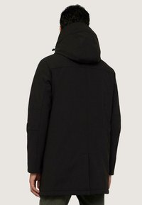 Chaqueta negra con capucha, de textura suave, con cierre de cremallera y bolsillos laterales. Presenta paneles cosidos y un corte ligeramente más largo en la parte de atrás.