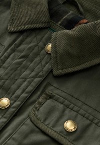 Grön vadderad jacka med ett kordflossakrage, metallknappar och diagonala sömmar. Inkluderar en ficka med strukturerade accenter.