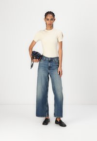 Κρέμ ζιβάγκο μπλουζάκι με κοντά μανίκια, ψηλόμεσα μπλε παντελόνια με φαρδύ πόδι και μαύρα λουστρίνια, κρατώντας μια σκούρα μπλε τσάντα με κορδόνια.