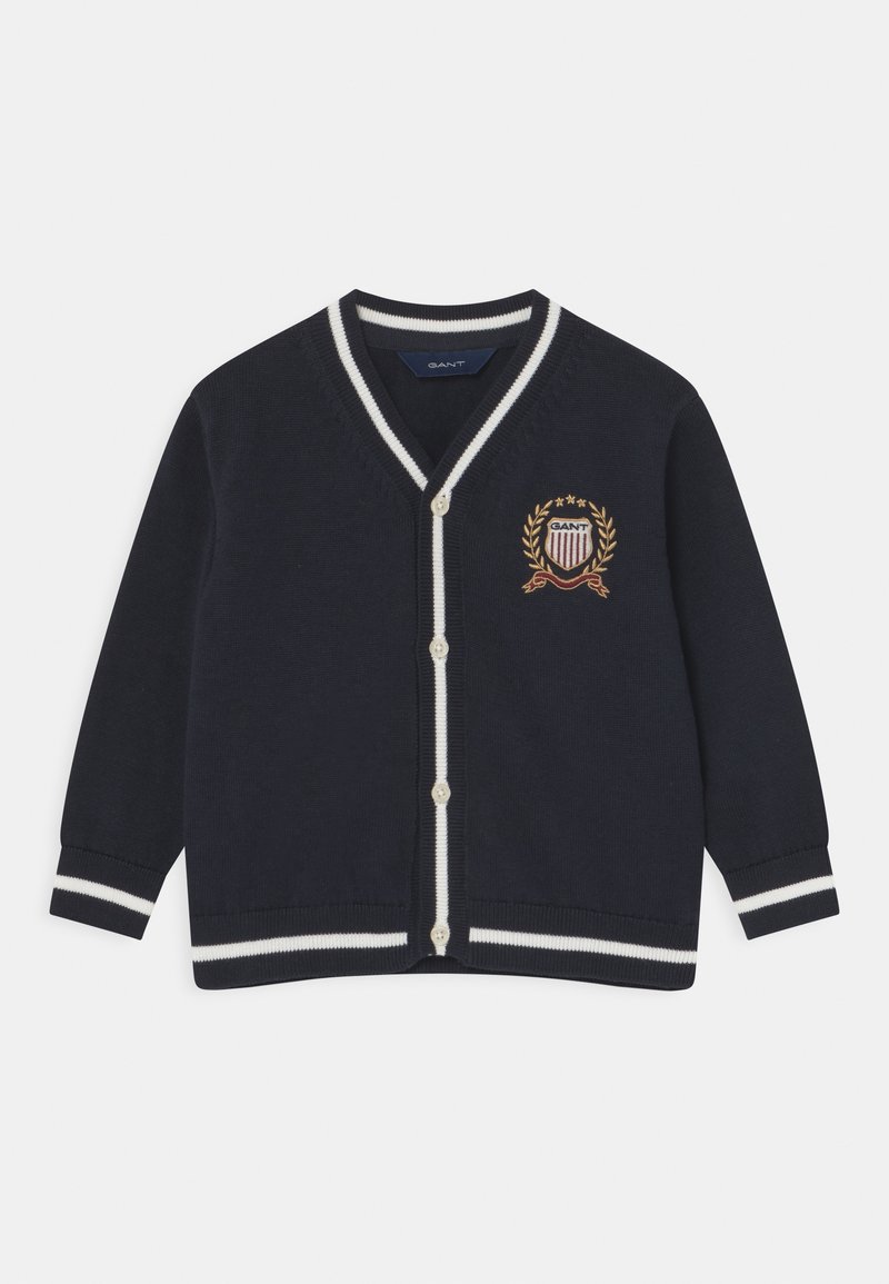 Námořnický svetr s bílými pruhy, ve tvaru V, se zapínáním na knoflíky. Obsahuje vyšitý emblém s vavřínem a štítovými detaily.