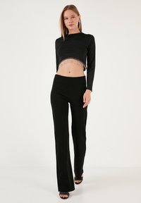 Čierny cropped top s dlhým rukávom a strapcovým lemom, doplnený o priliehavé čierne nohavice. Hladká tkanina, minimalistický dizajn, strih na mieru a topánky na otvorenej špičke.