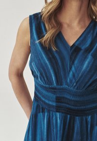 Blå ärmlös klänning med V-ringning, med en smockad midja och en mjuk, silkeslen textur. Mönster i varierande nyanser skapar en subtil gradation.