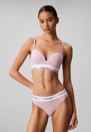 Jonge vrouw draagt lichtroze Calvin Klein beha en ondergoed set, staand met één hand op de heup tegen een effen grijze achtergrond.