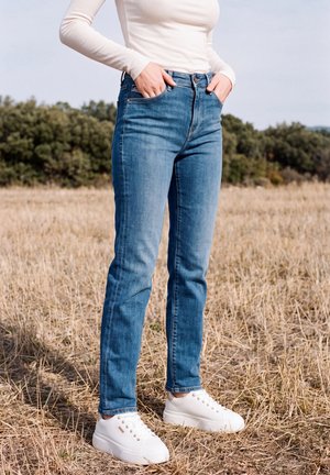 Une personne se tient dans un champ sec, vêtue d’un jean bleu, d’un haut blanc et de baskets blanches, les mains glissées dans les poches avant.