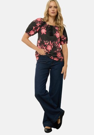 Donna con blusa nera a stampa floreale rosa, jeans larghi scuri e scarpe nere, in piedi con una mano in tasca.