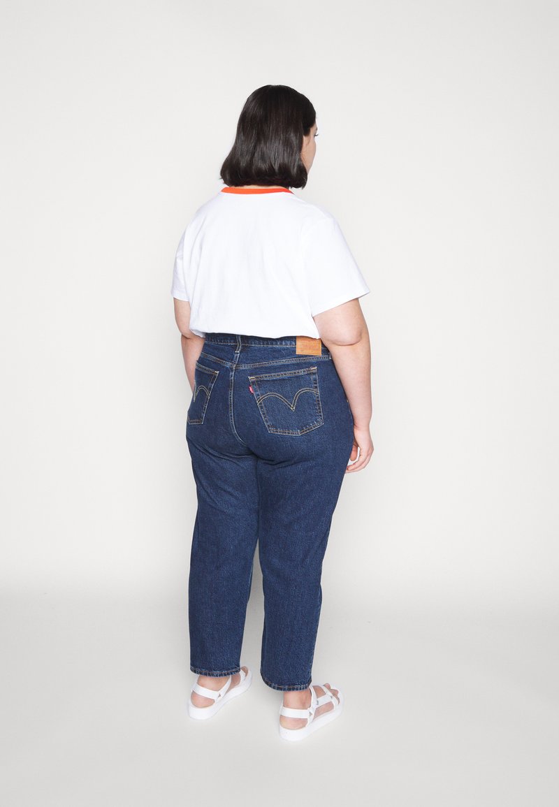 Hochtaillierte blaue Jeans aus Denim mit klassischem Fünf-Taschen-Design, kombiniert mit einem weißen T-Shirt mit orangefarbenem Kragen und weißen Sandalen.