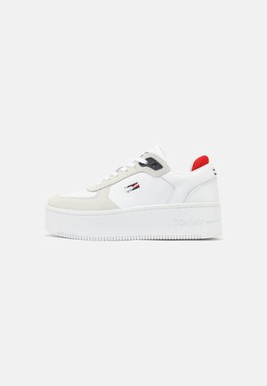 Biały sneakers na platformie z beżowymi akcentami, czerwoną pętelką na pięcie, czarnym oczkiem i małym logo flagi po boku, pokazany z boku na białym tle.