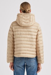 Beige Steppjacke mit Kapuze, mit gesteppte Abschnitte, weiten Ärmeln und kurzer Länge, kombiniert mit blauen Jeans.