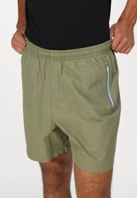 Bermudas caqui con cintura elástica y un bolsillo con cremallera lateral que presenta acentos en azul y verde. Fabricadas con una tela ligera y texturizada.
