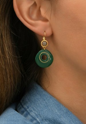 Les boucles d'oreilles en or en forme de créoles présentent un pendentif rond en résine verte avec une découpe circulaire et des accents dorés texturés.