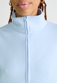 Svetlo modra zip-up pulover s ovratnikom z gladko teksturo in kontrastnim zadrgo, ki ima šivane šive in visok zgornji del.