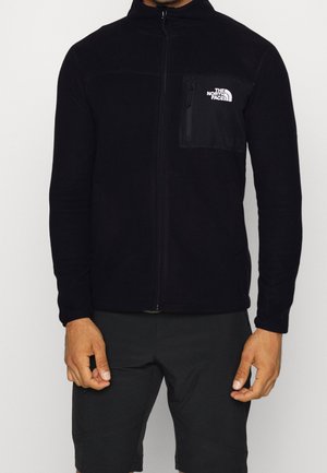 Muž nosící černou fleecovou bundu na zip s logem The North Face na levé straně hrudi a černé šortky na jednobarevném pozadí.