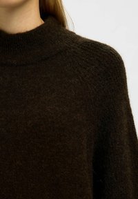 Tmavě hnědý pletený rolák s vysokým límcem a strukturovaným povrchem, který má volný střih a měkký, huňatý materiál.