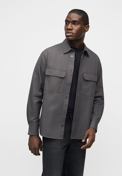 Άνδρας που φορά σκούρο γκρι πουκάμισο με κουμπιά και δύο τσέπες στο στήθος πάνω από μαύρο πουκάμισο και σκούρο τζιν, κοιτάζοντας προς τα πλάγια ενάντια σε απλό φόντο.