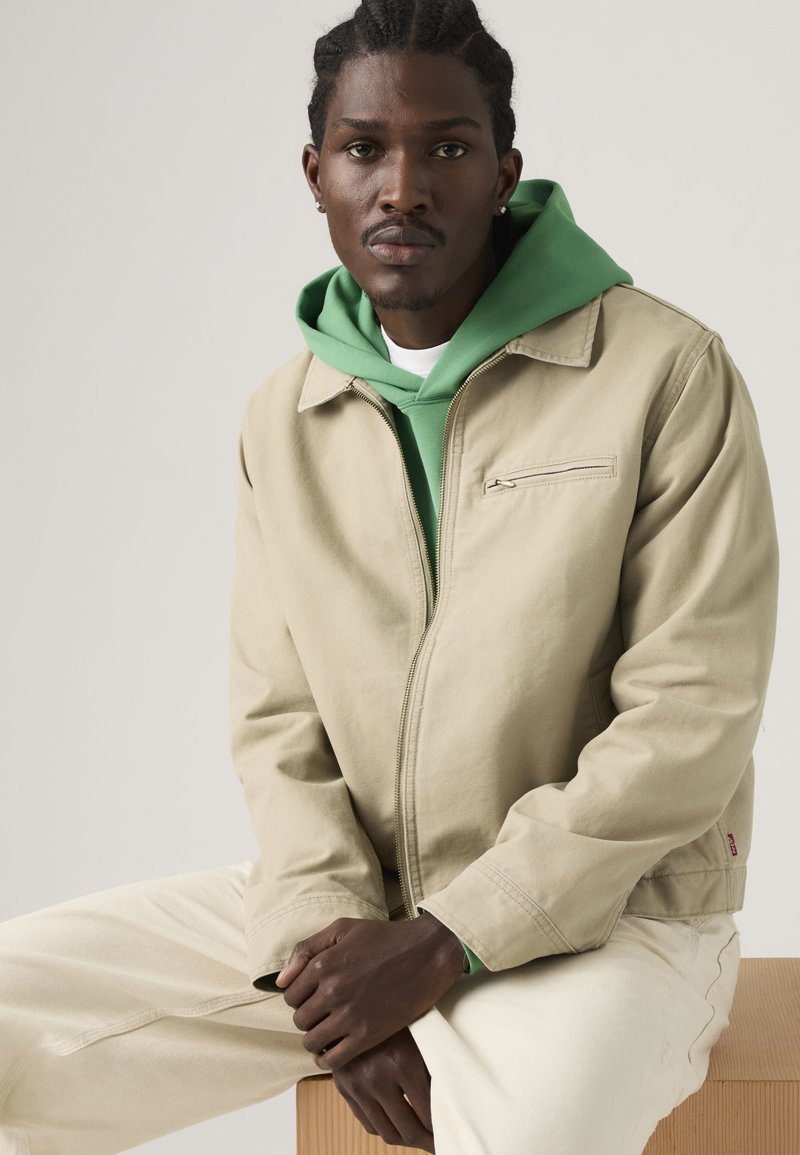 Mann mit geflochtenem Haar sitzt, trägt eine beige Jacke über einem grünen Hoodie und hellfarbige Hose vor einfachem hellem Hintergrund.