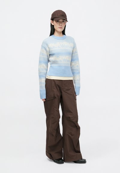 Šviesiai mėlynas megztinis su logotipo raštu, guminiais rankogaliais; rudi plačiakulkiai kelnes; juodi batai; rudas kepurė. Lygūs audiniai, kasdieniškas siluetas.