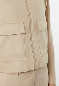 Beżowa kurtka zapinana na zamek wykonana z miękkiego materiału, wyposażona w dwie przednie naszywane kieszenie, złoty zamek błyskawiczny i dopasowany krój. Gładka tekstura.