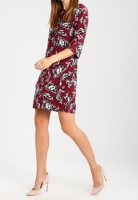 Robe bordeaux à motifs floraux et de feuilles blancs, en tissu léger, dotée de manches 3/4 et d'une coupe au-dessus du genou. Associée à des talons hauts nude.