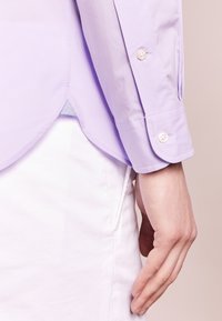 Lila skjorta med knappar, rundad nederkant, långa ärmar och vita knappdetaljer. Slät textur, i kombination med vitt material nedanför.