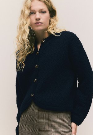 Cardigan texturat de culoare bleu marin cu guler rotund și nasturi mari, asortat cu o fustă în carouri bej și maro. Material neted, croială lejeră.