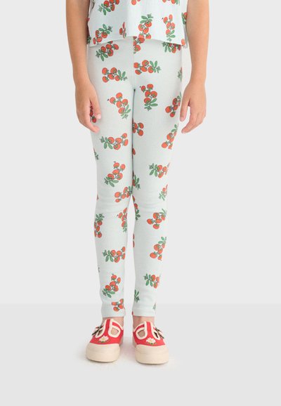 Enfant portant des leggings bleu clair avec des motifs de cerises rouges et des chaussures red slip-on décorées de fleurs, debout devant un fond blanc.