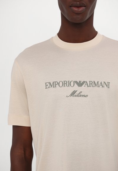 Šviesiai ruda marškinėlių suapvalintu kaklu, pagaminti iš lengvos medžiagos. Turi trumpas rankoves ir tamsiai žaliai spausdinta "EMPORIO ARMANI Milano".