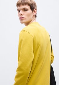 Κίτρινο βαμβακερό μακρυμάνικο πουκάμισο με στρογγυλή λαιμόκοψη, εμφανιζόμενο από πίσω, σε συνδυασμό με μια μαύρη τσάντα ώμου. Απαλή υφή, casual εφαρμογή.