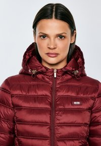 Žena s rovnými tmavými vlasy, oblečená ve vínově červené prošívané bundě na zip s kapucí a kovovými šňůrkami s brzdičkami, čelem k nám na světlém pozadí.