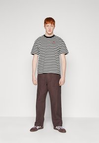 T-shirt rayé noir et blanc avec un logo rouge sur la poitrine, associé à un pantalon marron ample et des chaussures foncées. Design simple et décontracté.