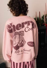 Różowy sweatshirt z vintage grafiką wiśni i tekstem. Projekt zawiera kokardę oraz dodatkowe frazy w kontrastującym ciemniejszym odcieniu.