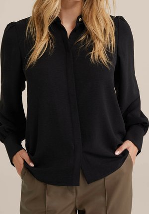 Blond vrouw die een zwarte blouse met lange mouwen en kraag draagt, dichtgeknoopt, en olijfgroene broek, handen in de zakken.