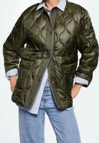 Khaki prošívaná bunda s límečkem, předními knoflíky a dvěma předními kapsami. Nesená přes světle modrou košili a volné džíny.