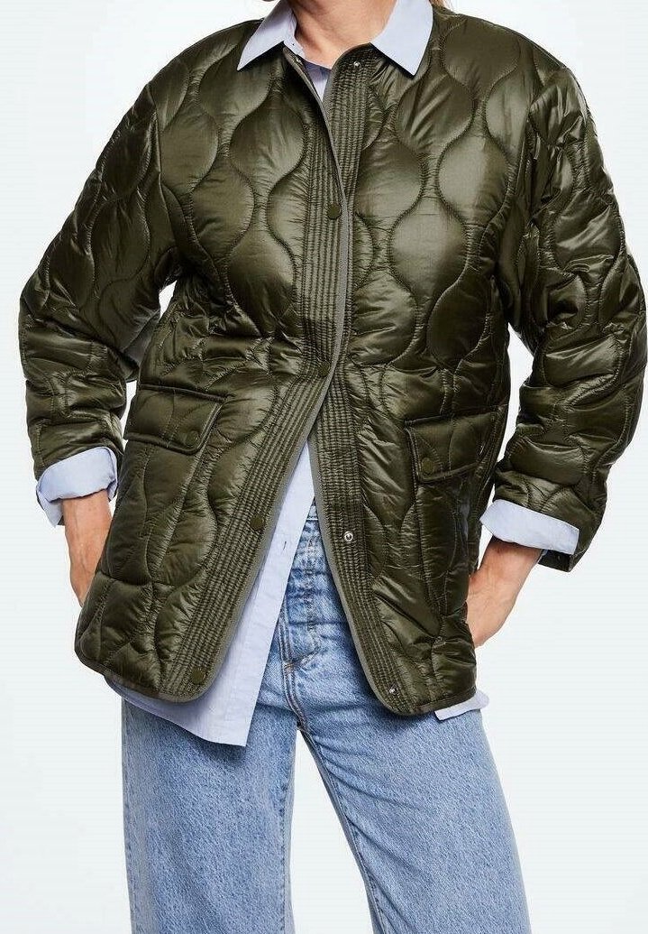 Khaki prošívaná bunda s límečkem, předními knoflíky a dvěma předními kapsami. Nesená přes světle modrou košili a volné džíny.