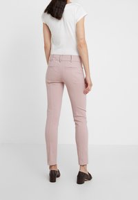 Vrouw die met de rug naar voren staat, gekleed in lichtroze strakke broek, witte korte mouw top en zwarte hoge hakken tegen een effen achtergrond.