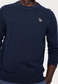 Mörkblå stickad tröja med rundad hals och gröna ribbade detaljer. Har en liten, flerfärgad hästlogga på bröstet.