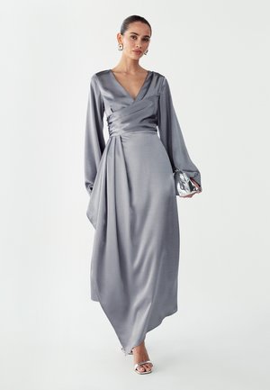 Vestido largo de satén gris con diseño cruzado, escote en V profundo y mangas largas; presenta tela drapeada y una silueta fluida. Clutch plateado a juego.