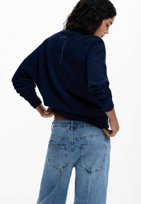 Femeie ajustând blugi bleu deschis, purtând un hanorac bleumarin cu detalii vizibile de cusături pe partea superioară a spatelui, văzută din profil pe un fundal alb.