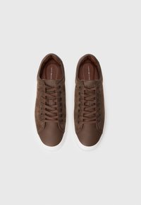 Barna nubuk sportcipők sima barna bőrből készült orrával, fekete fűzőlyukakkal és fehér gumi talppal. A fűzők barna hurkokon keresztül vannak fűzve.