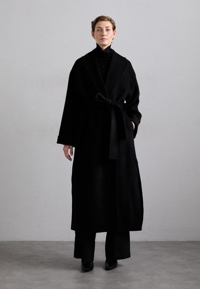 Langer schwarzer Mantel mit Bindegürtel, aus weichem Material, mit weiten Ärmeln und minimalistischer Gestaltung, getragen über schwarzen Hose und Schuhen.