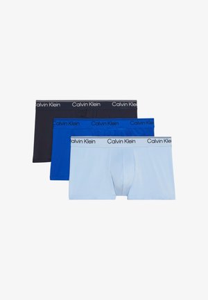 Homme musclé debout pieds nus portant un boxer Calvin Klein bleu clair sur un fond gris uni.