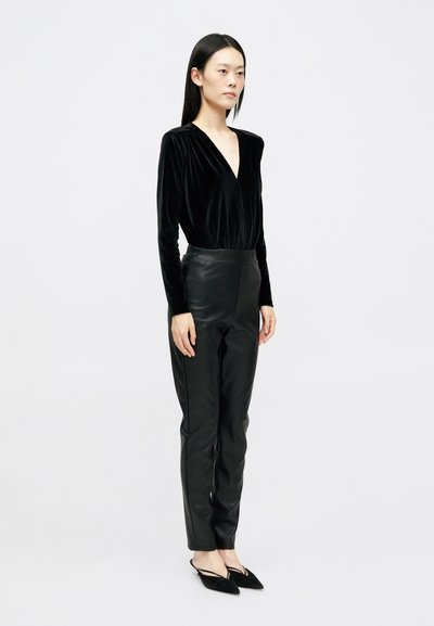 Femeie îmbrăcată cu un top negru din catifea cu decolteu în V, pantaloni negri din piele și pantofi slingback negri cu vârf ascuțit, stând în fața unui fundal alb simplu.