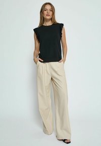 Haut à manches ondulées noires associé à un pantalon large beige. Tissu lisse, coupe décontractée, poches latérales, détails minimaux et chaussures visibles.