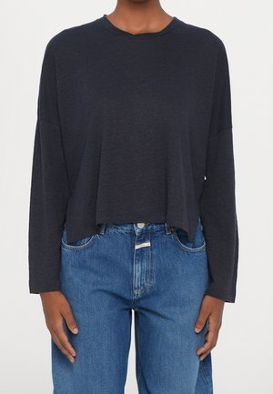 Personne portant un haut bleu marine foncé à manches longues amples et un jean en denim bleu taille haute sur un fond uni.