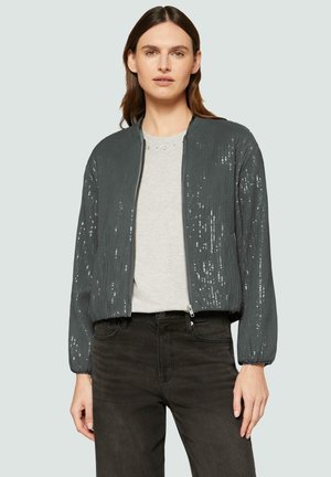 Veste grise pailletée avec fermeture à glissière à l'avant, poignets côtelés et coupe décontractée. Portée par-dessus un haut gris clair et associée à un jean foncé.